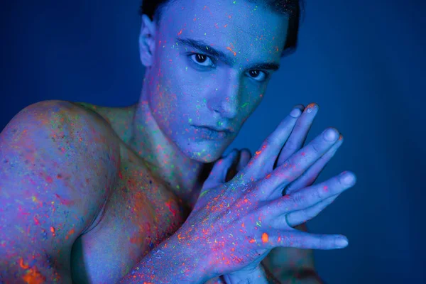 Homme jeune, expressif et torse nu en peinture corporelle rayonnante et colorée posant avec les mains jointes et regardant la caméra sur fond bleu avec un effet d'éclairage cyan — Photo de stock