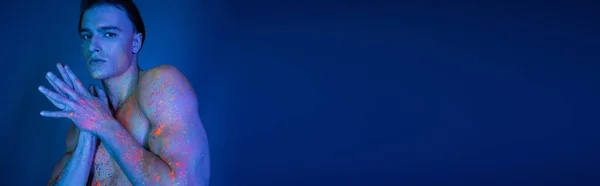 Hombre joven, carismático y sin camisa con torso muscular posando en colorida pintura de cuerpo de neón mientras mira a la cámara sobre fondo azul con efecto de iluminación cyan, pancarta — Stock Photo