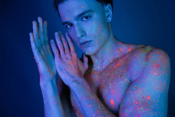 Youthful, self-assured and shirtless man in vibrant and colorful neon body paint holding hands near face while looking at camera on blue background with cyan lighting effect — Stock Photo