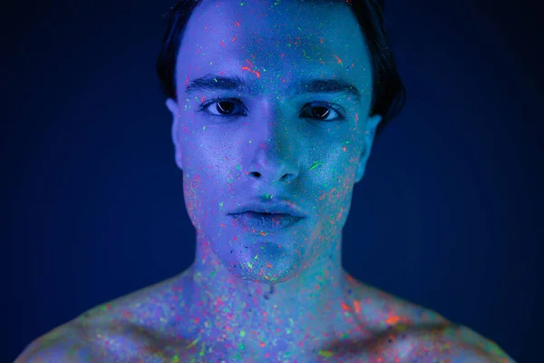 Portrait of confident, self-assured man with bare shoulders posing in vibrant and colorful neon body paint while looking at camera on blue background with cyan lighting effect — Stock Photo