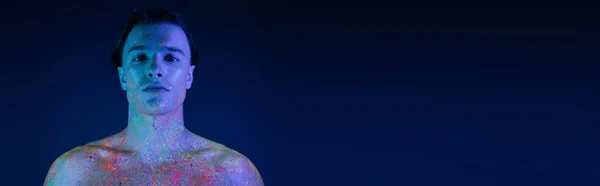 Ritratto di giovane uomo bello e sicuro di sé guardando la macchina fotografica mentre posa in vernice luminosa e colorata del corpo al neon su sfondo blu con effetto di luce ciano, banner — Foto stock