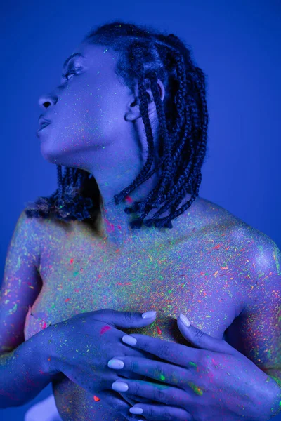Jovem, sensual e nu-peito mulher americana africana com dreadlocks cobrindo o peito com as mãos enquanto posando em tinta corporal de néon colorido no fundo azul com efeito de iluminação ciano — Fotografia de Stock