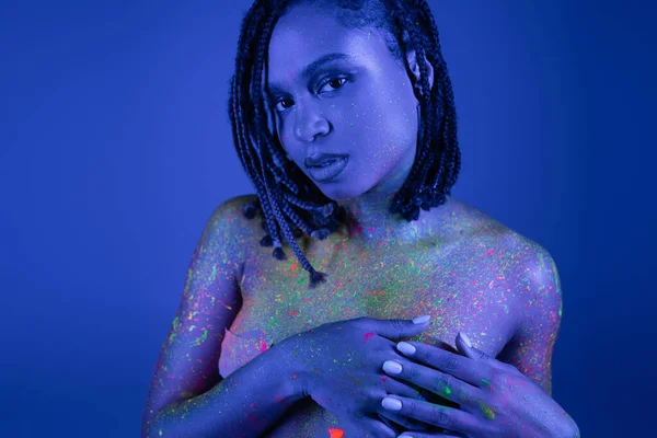 Mujer afroamericana desnuda y provocativa con rastas posando en pintura corporal de neón multicolor, cubriendo el pecho con las manos y mirando a la cámara sobre fondo azul con efecto de iluminación cian - foto de stock