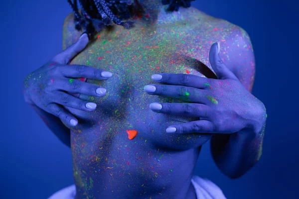 Partial view of youthful and bare-chested african american woman in vibrant and colorful neon body paint covering breast with hands on blue background with cyan lighting effect — Stock Photo