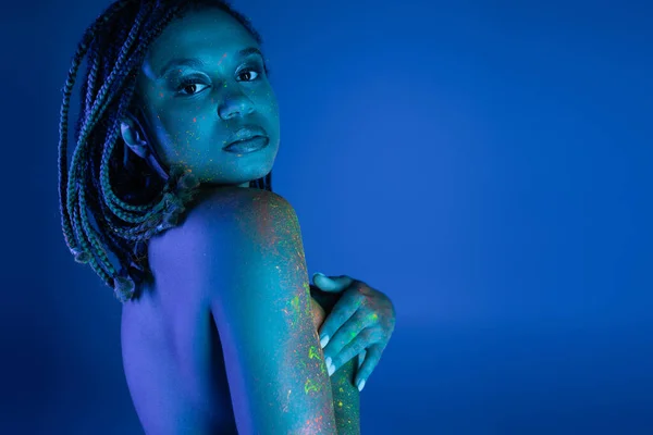 Tentadora mujer afroamericana con rastas cubriendo el pecho con las manos y mirando a la cámara mientras posa en colorida pintura corporal de neón sobre fondo azul con efecto de iluminación cian - foto de stock
