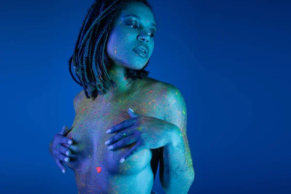 Charming and nude african american woman posing in radiant and colorful neon body paint while looking away and covering breast with hands on blue background with cyan lighting effect — Stock Photo