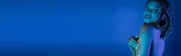 Співчутлива і гола афроамериканка з дредлоками, що покривають груди руками, позуючи в барвистій неоновій фарбі тіла на синьому фоні з ефектом блакитного освітлення, банер — стокове фото