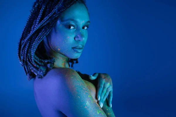 Интригующая африканская американка в разноцветной неоновой краске для тела смотрит в камеру и закрывает грудь руками, позируя на синем фоне с эффектом синего освещения — стоковое фото