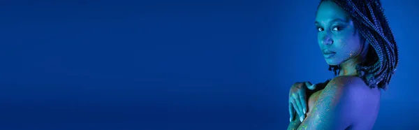 Молодая, соблазнительная и обнаженная грудь африканской женщины в красочной неоновой краске для тела, смотрящей на камеру и прикрывающей грудь рукой на голубом фоне с эффектом синего освещения, баннер — стоковое фото