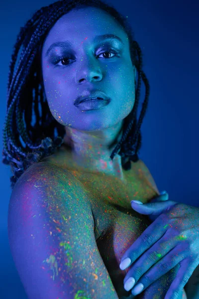 Expressive and charming african american woman in radiant and multicolored neon body paint covering breast with hands and looking at camera on blue background with cyan lighting effect — Stock Photo