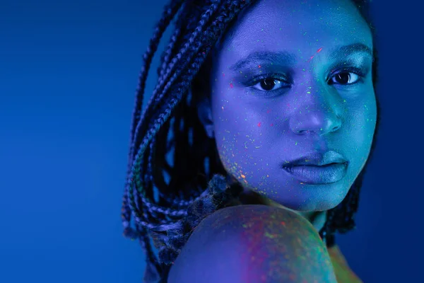 Portrait of youthful and intriguing african american woman with dreadlocks, in colorful neon body paint looking at camera on blue background with cyan lighting effect — Stock Photo