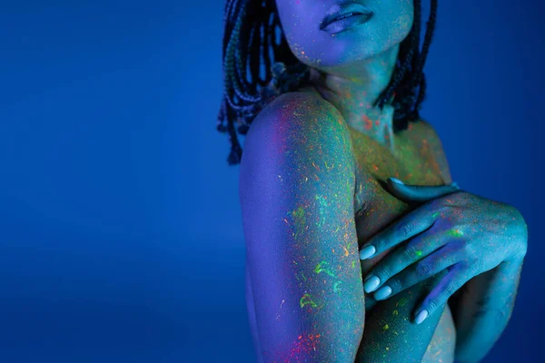 Partial view of nude african american woman in bright and colorful neon body paint covering breast with hands while standing on blue background with cyan lighting effect — Stock Photo