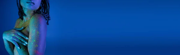 Vista parziale di giovane donna afroamericana con dreadlocks posa in vernice colorata al neon corpo su sfondo blu con effetto luce ciano, banner — Foto stock