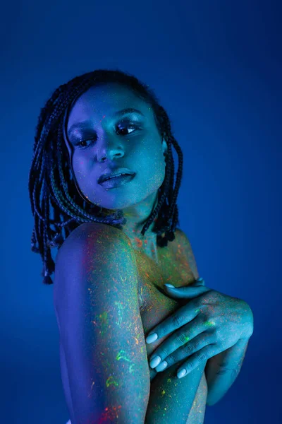 Jovem e tentadora mulher afro-americana nua de peito com dreadlocks, em tinta corporal de néon colorido, cobrindo o peito com as mãos e olhando para o fundo azul com efeito de iluminação ciano — Fotografia de Stock