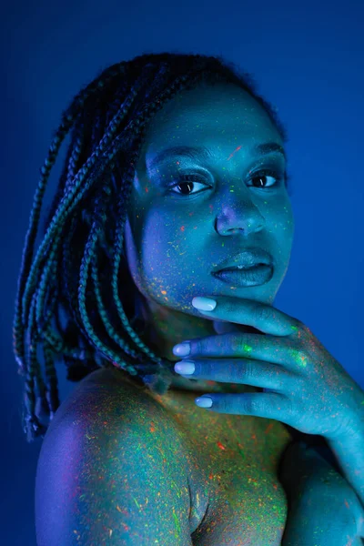Portrait of youthful and eye-catching african american woman in multicolored neon body paint holding hand near face and looking at camera on blue background with cyan lighting effect — Stock Photo