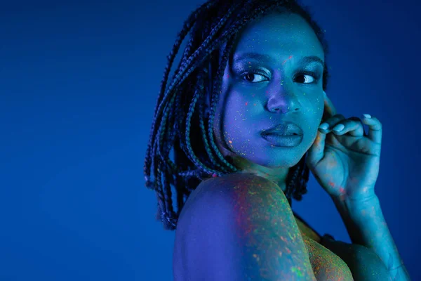 Молодая и привлекательная африканская американка в разноцветной неоновой краске для тела, держась за руку рядом с лицом, отводя взгляд на синий фон с эффектом синего освещения — стоковое фото