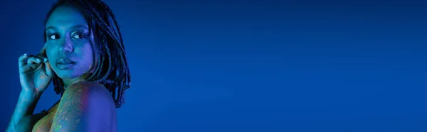 Femme américaine africaine sensuelle avec dreadlocks et épaule nue, dans la peinture au néon colorée rayonnante du corps regardant loin sur fond bleu avec effet d'éclairage cyan, bannière — Photo de stock