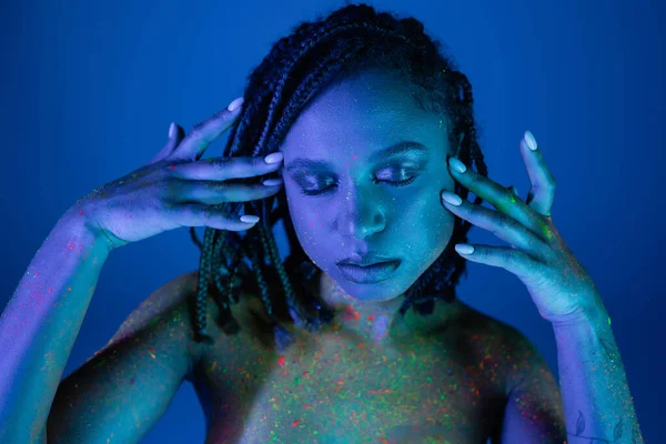 Sedutor e nu-peito mulher americana africana em colorido neon pintura corporal posando com olhos fechados e mãos perto do rosto no fundo azul com efeito de iluminação ciano — Fotografia de Stock