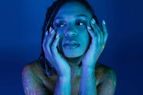 Verführerische afrikanisch-amerikanische Frau mit Dreadlocks, die die Hände vor dem Gesicht hält, während sie in bunter Neon-Körperfarbe posiert und vor blauem Hintergrund mit Cyan-Lichteffekt wegschaut — Stockfoto