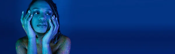 Retrato de mujer afroamericana joven con rastas, en colorida pintura de cuerpo de neón, cogidas de la mano cerca de la cara y mirando hacia otro lado sobre fondo azul con efecto de iluminación cian, pancarta - foto de stock