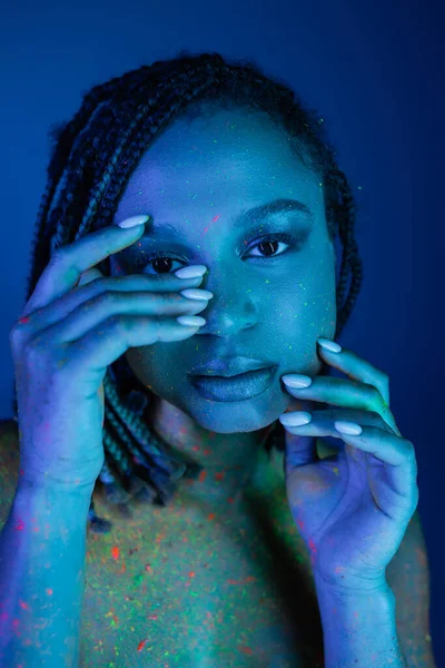 Retrato de jovem e sedutora mulher afro-americana com dreadlocks segurando as mãos perto do rosto enquanto posando em tinta corporal de néon colorido em fundo azul com efeito de iluminação ciano — Fotografia de Stock