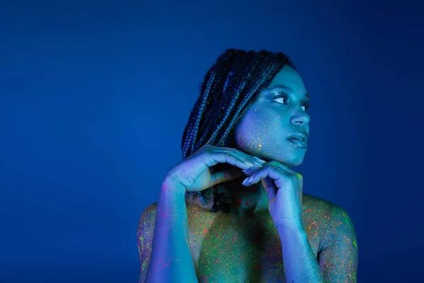 Joven y cautivadora mujer afroamericana con rastas, en pintura de cuerpo de neón brillante y colorido cogidas de la mano cerca de la barbilla y mirando hacia otro lado sobre fondo azul con efecto de iluminación cian - foto de stock