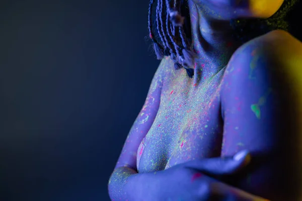 Visão parcial da mulher americana africana jovem e nua com dreadlocks posando em tinta corporal de néon colorido e cobrindo o peito com as mãos no fundo azul com efeito de iluminação amarela — Fotografia de Stock