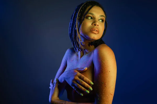 Mulher afro-americana nua-peito com dreadlocks de pé na pintura do corpo de néon colorido, cobrindo o peito com as mãos e olhando para longe no fundo azul com efeito de iluminação amarela — Fotografia de Stock