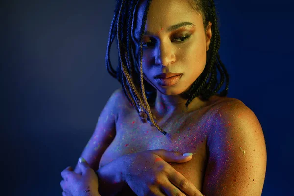 Appealing african american woman with dreadlocks covering breast with hands while posing in radiant and colorful neon body paint on blue background with yellow lighting effect — Stock Photo