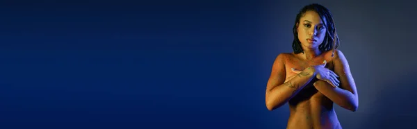 Tentante et nue femme afro-américaine dans coloré néon corps peinture couvrant poitrine avec les mains tout en regardant la caméra sur fond bleu avec effet d'éclairage jaune, bannière — Photo de stock