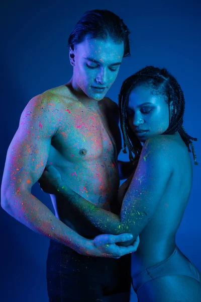 Intimer Moment eines gemischtrassigen Paares in bunter Neon-Körperfarbe, das sich auf blauem Hintergrund mit Cyanbeleuchtung umarmt, Mann mit muskulösem Oberkörper und hemdlose Afroamerikanerin mit Dreadlocks — Stockfoto