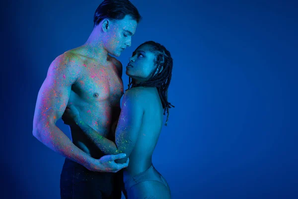 Sexy pareja multicultural en colorida pintura de cuerpo de neón mirándose el uno al otro sobre fondo azul con iluminación cyan, hombre sin camisa con cuerpo muscular y atractiva mujer afroamericana - foto de stock