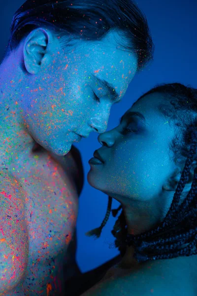 Impassioned interracial couple standing face to face with closed eyes, bare-chested man and african american woman with dreadlocks on blue background with cyan lighting — Stock Photo