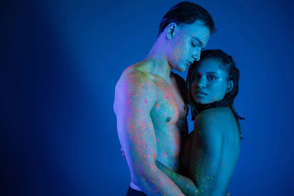 Jeune femme afro-américaine avec dreadlocks, en peinture au néon coloré, embrassant homme torse nu sexy avec corps musclé et regardant la caméra sur fond bleu avec éclairage cyan — Photo de stock