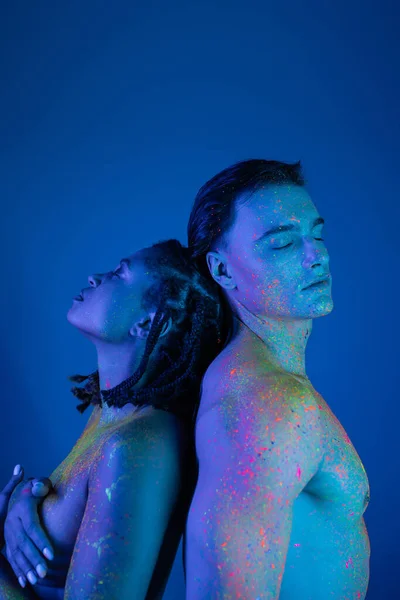 Junge interrassische Paar in bunten Neon-Körperfarbe Rücken an Rücken mit geschlossenen Augen, nackte afrikanisch-amerikanische Frau bedeckt Brust in der Nähe muskulösen Mannes auf blauem Hintergrund mit Cyan-Beleuchtung — Stockfoto