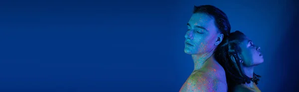 Young interracial couple in colorful neon body paint, with bare shoulders and closed eyes, standing back to back on blue background with cyan lighting, banner — Stock Photo