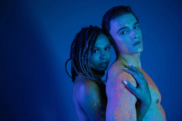 Sexy couple interracial dans coloré néon corps peinture regardant caméra sur fond bleu avec éclairage cyan, nu afro-américaine femme embrassant homme torse nu avec corps musclé — Photo de stock