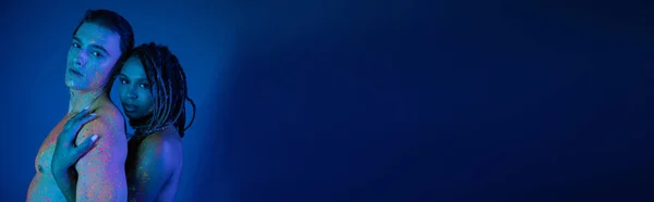 Romantisches Paar in bunter Neon-Körperbemalung mit Blick in die Kamera auf blauem Hintergrund mit Cyanbeleuchtung, afrikanisch-amerikanische Frau mit Dreadlocks umarmt sexy hemdlosen Mann mit muskulösem Oberkörper, Banner — Stockfoto