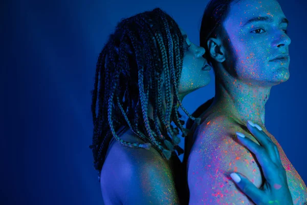 Momento intimo di giovane coppia interrazziale in colorato vernice corpo al neon su sfondo blu con illuminazione ciano, appassionato donna afro-americana abbracciando uomo carismatico con spalle nude — Foto stock
