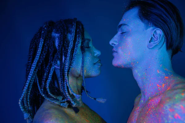 Side view of interracial couple with bare shoulders standing face to face with closed eyes, handsome man and african american with dreadlocks on blue background with cyan lighting — Stock Photo
