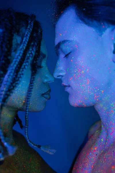 Bare-chested interracial couple in colorful neon body paint standing face to face with closed eyes, youthful man and african american woman with dreadlocks on blue background with cyan lighting — Stock Photo