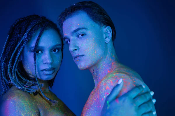 Youthful multicultural couple with bare shoulders, in colorful neon body paint looking at camera on blue background with cyan lighting, charismatic man and african american woman with dreadlocks — Stock Photo