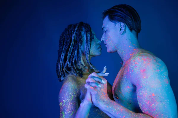 Side view of interracial couple in colorful neon body paint standing with clenched hands, african american woman with dreadlocks and shirtless muscular man on blue background with cyan lighting — Stock Photo