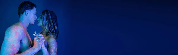 Hombre musculoso sin camisa y mujer afroamericana con rastas mirándose el uno al otro y de pie con las manos apretadas en colorida pintura de cuerpo de neón sobre fondo azul con iluminación cian, pancarta - foto de stock