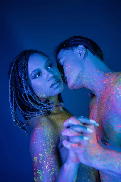 Expressive interracial couple in colorful neon body paint standing with clenched hands on blue background with cyan lighting, sexy shirtless man kissing african american woman with dreadlocks — Stock Photo