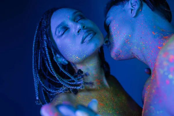 Sexy interracial couple in colorful neon body paint, african american woman with dreadlocks and closed eyes near young bare-chested man on blue background with cyan lighting — Stock Photo