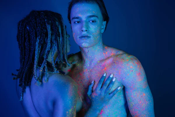 Young and nude interracial couple in colorful neon body paint, african american woman with dreadlocks near shirtless man with muscular body on blue background with cyan lighting — Stock Photo
