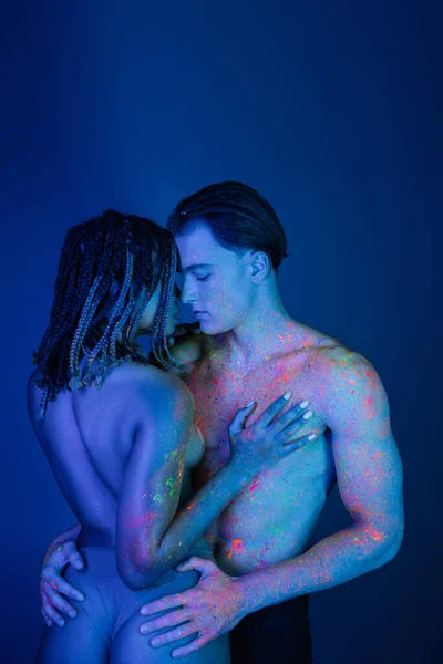 Momento íntimo de casal interracial em tinta corporal de néon colorido, homem sem camisa confiante abraçando nádegas sensuais de jovem mulher americana africana no fundo azul com iluminação ciana — Fotografia de Stock