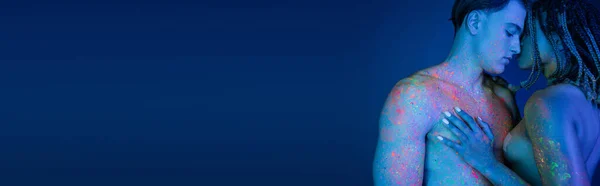 Momento íntimo de casal interracial em tinta corporal de néon colorido no fundo azul com iluminação ciana, mulher americana africana tocando no peito muscular do homem sem camisa, banner — Fotografia de Stock