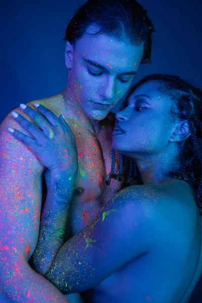 Couple multiculturel romantique en peinture corporelle colorée embrassant sur fond bleu avec éclairage cyan, femme africaine américaine nue avec dreadlocks et homme torse nu avec corps musclé — Photo de stock